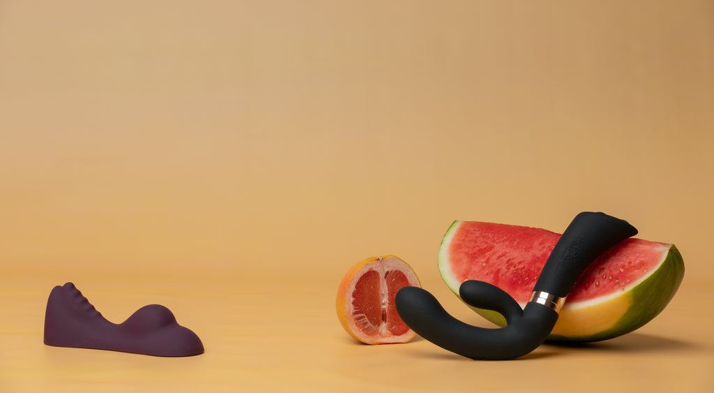 Image of the Ruby Glow on the left and the Enigma vibrator on the right. The Enigma is resting on a watermelon with a grapefruit sitting behind it. The background is orange 