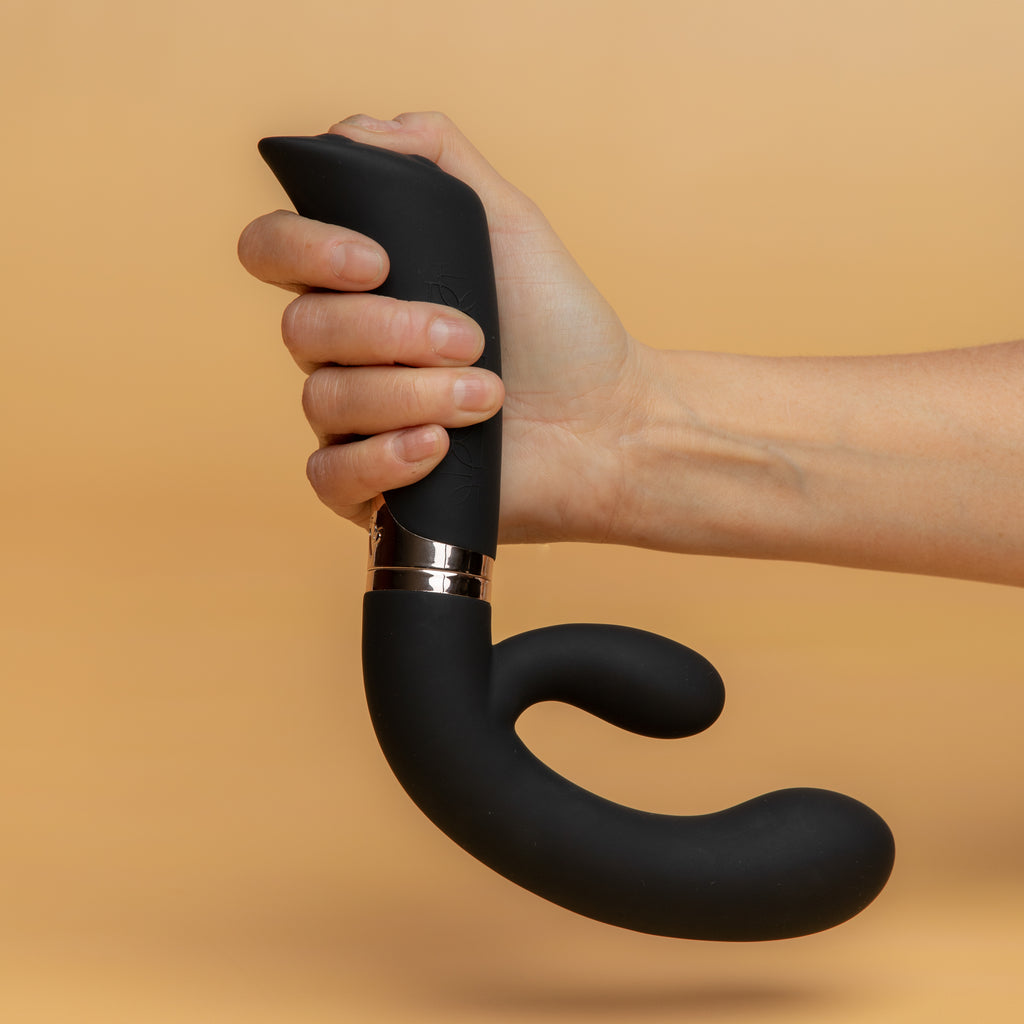 A hand holding the enigma with their fingers on the buttons. The bottom insertable part is slightly larger than the clitoral stimulator part. The toy is black. xes products