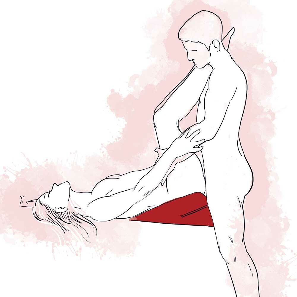 A woman lying with her back and bottom raised on the wedge. The partner is standing if front of the wedge holding on to the partners raised legs. 