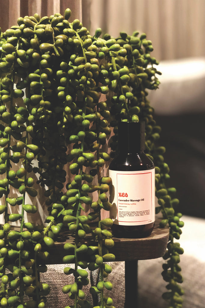Image of the Lavender Massage Oil enriched with magnesium sitting next to a plant. Behind the plan you can see curtains creating a cozy feeling. 